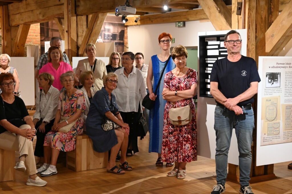 Uczestnicy finisażu pozują do wspólnego zdjęcia z prowadzącym; grupa osób stoi w sali wystawowej między planszami informacyjnymi, w tle widoczne są elementy ekspozycji (kliknięcie powiększy zdjęcie).