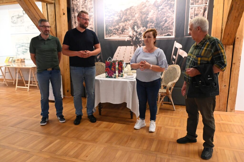 Cztery osoby stoją na przy stoliku nakrytym obrusem i witają gości; w tle znajduje się stół z materiałami oraz fotografie związane z wystawą (kliknięcie powiększy zdjęcie).
