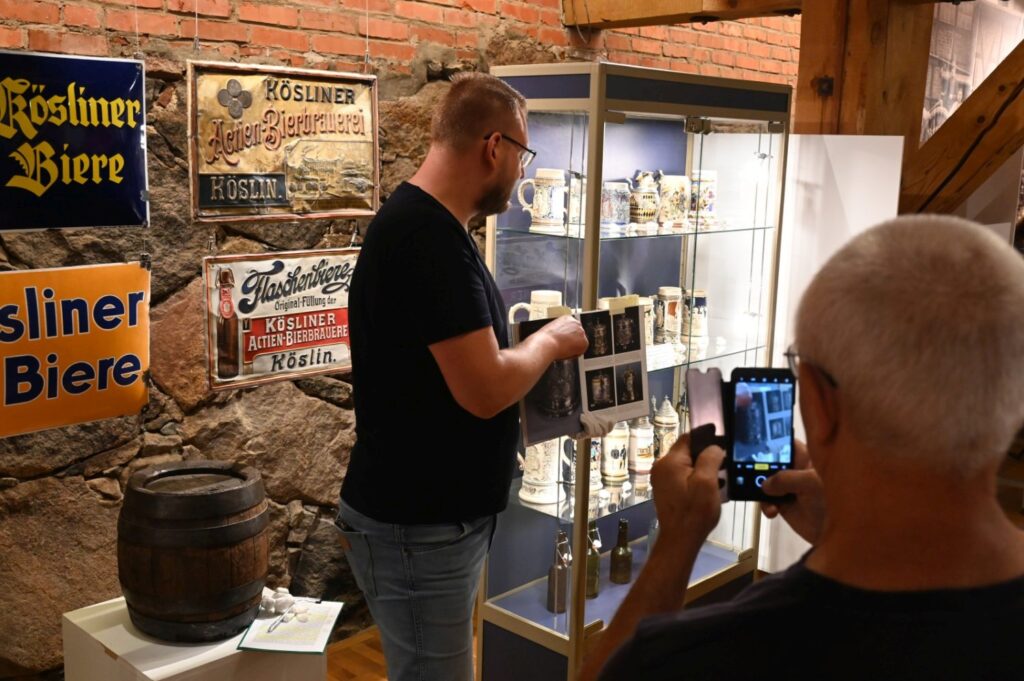 Kurator wystawy pokazuje i omawia eksponaty prezentowane w gablocie; w tle widoczne są historyczne reklamy piwa, beczka oraz kolekcja butelek i naczyń; na pierwszym planie jeden z uczestników fotografuje gablotę (kliknięcie powiększy zdjęcie).