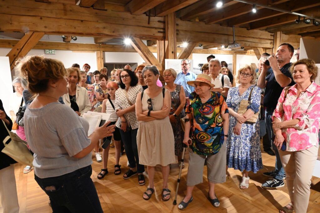 Uczestnicy finisażu słuchają prowadzącej; liczna grupa osób stoi w sali wystawowej, a w tle widoczne są drewniane belki oraz wielkoformatowa fotografia (kliknięcie powiększy zdjęcie).