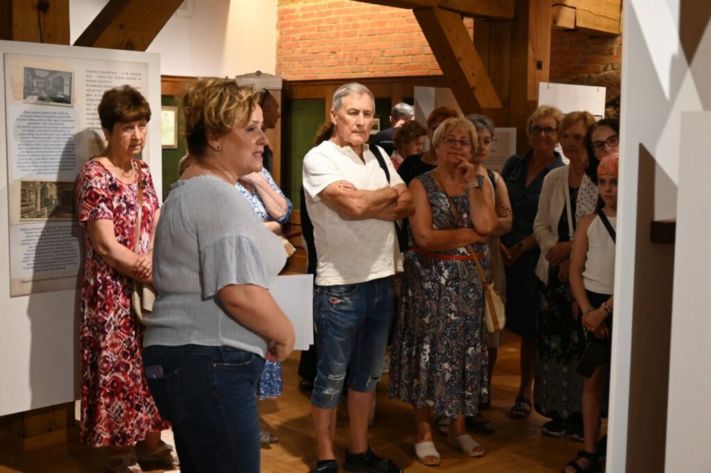 Prowadząca opowiada o ekspozycji podczas finisażu; grupa uczestników stoi w sali między planszami wystawowymi i słucha prezentacji (kliknięcie powiększy zdjęcie).