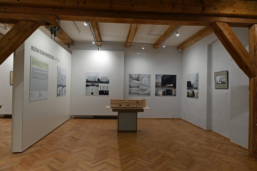 Centralna część sali wystawowej z planszami i fotografiami architektury; na środku ustawiono drewnianą makietę na podwyższeniu; ekspozycję otaczają białe ścianki działowe i drewniane elementy konstrukcji budynku (kliknięcie powiększy zdjęcie).