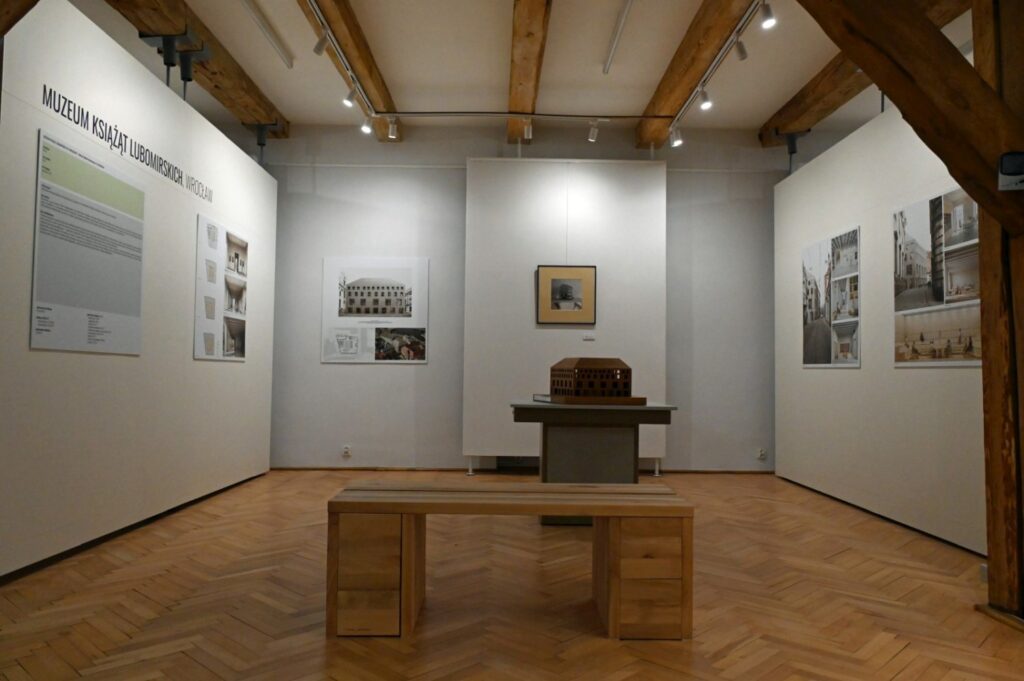 Sala wystawowa z planszami informacyjnymi i fotografiami architektury; pośrodku znajduje się makieta budynku na podwyższeniu; przed nią ustawiono drewnianą ławkę (kliknięcie powiększy zdjęcie).