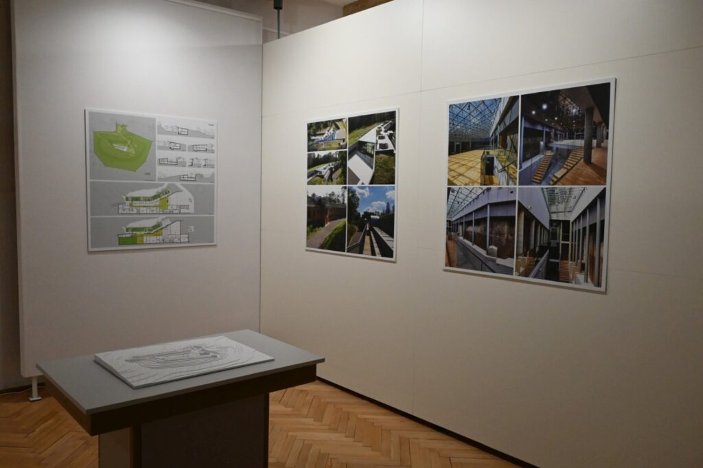 Zbliżenie na fragment ekspozycji architektonicznej; na białych ścianach znajdują się kolorowe plansze, schematy i fotografie budynków oraz przestrzeni miejskich; przed ścianą ustawiono niski stół z materiałem ekspozycyjnym (kliknięcie powiększy zdjęcie).