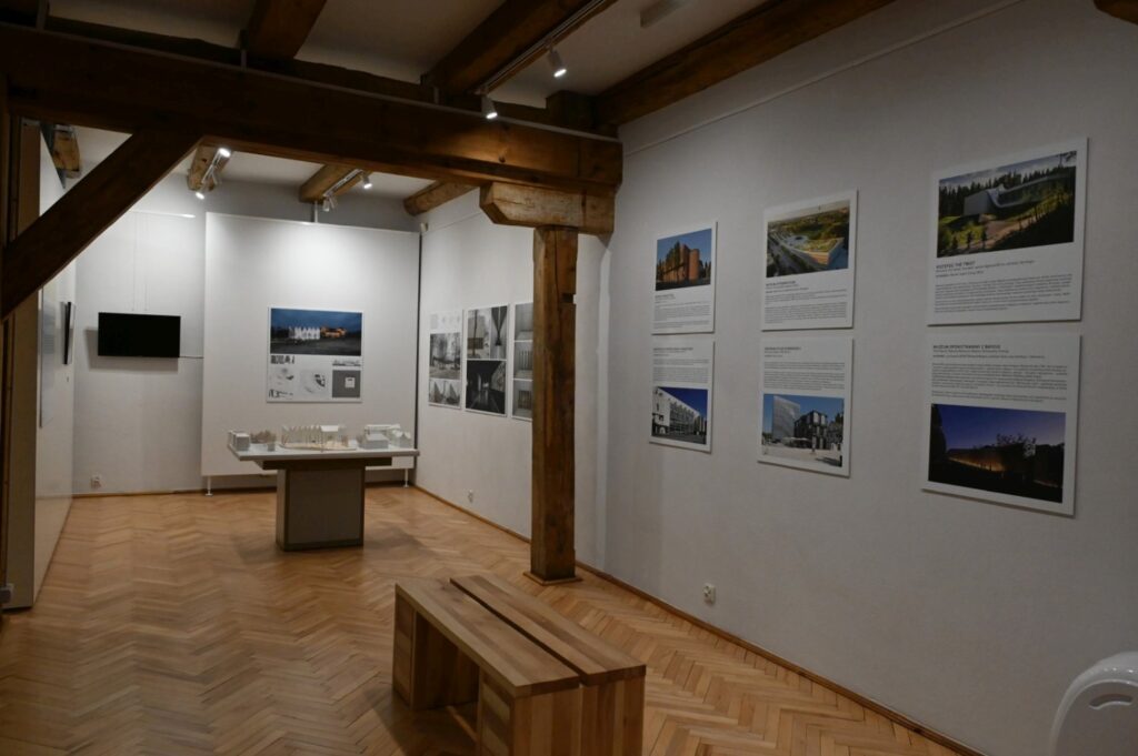 Widok kolejnej części wystawy architektonicznej; na ścianach rozmieszczono plansze z tekstami i fotografiami budynków; pośrodku ustawiono stół ekspozycyjny z makietą; w tle widoczna jest ławka dla zwiedzających (kliknięcie powiększy zdjęcie).