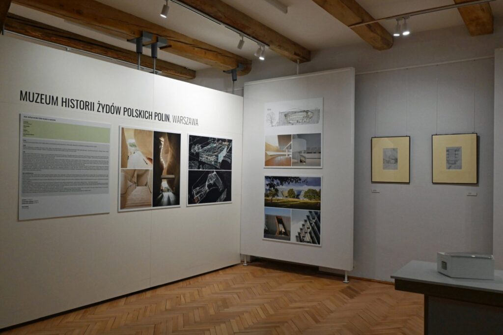 Widok wystawy poświęconej historii i architekturze; na ścianie znajduje się napis „MUZEUM HISTORII ŻYDÓW POLSKICH POLIN, WARSZAWA”; obok umieszczono fotografie i plansze informacyjne; na pierwszym planie znajduje się stół ekspozycyjny (kliknięcie powiększy zdjęcie).