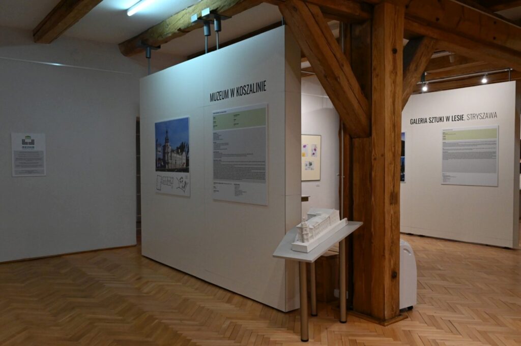 Widok sali wystawowej z drewnianymi belkami stropowymi i ekspozycją plansz oraz makiet; na ścianach prezentowane są informacje dotyczące architektury; na pierwszym planie stoi niewielki stolik z makietą (kliknięcie powiększy zdjęcie).