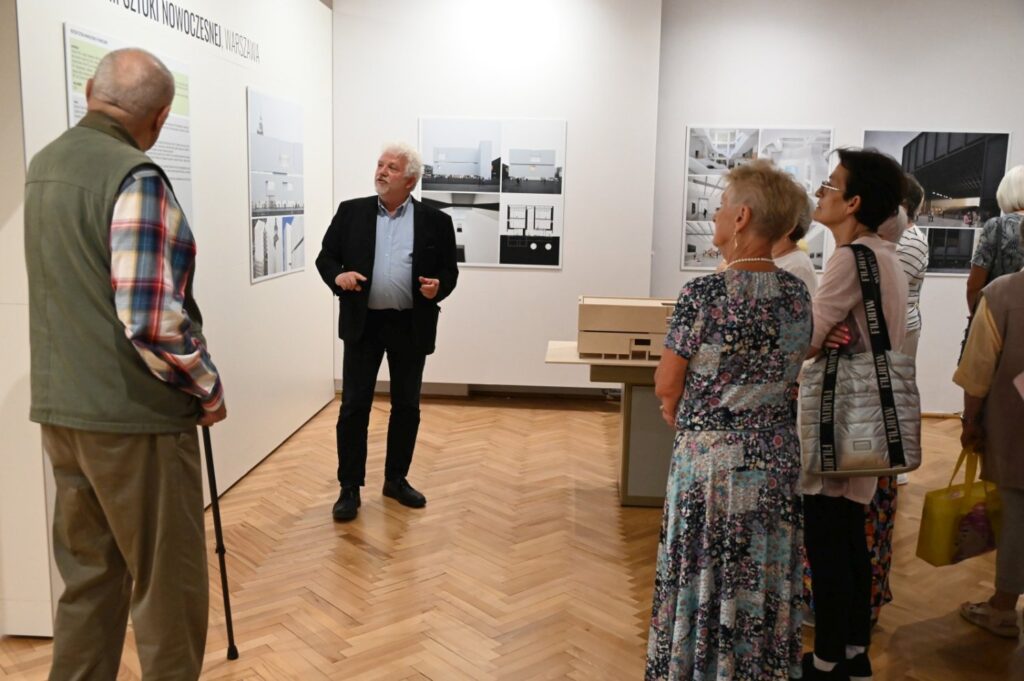 Oprowadzanie po wystawie w muzealnej sali; mężczyzna pokazuje grupie zwiedzających plansze i fotografie na ścianach; na środku sali znajduje się drewniany stół z makietą (kliknięcie powiększy zdjęcie).