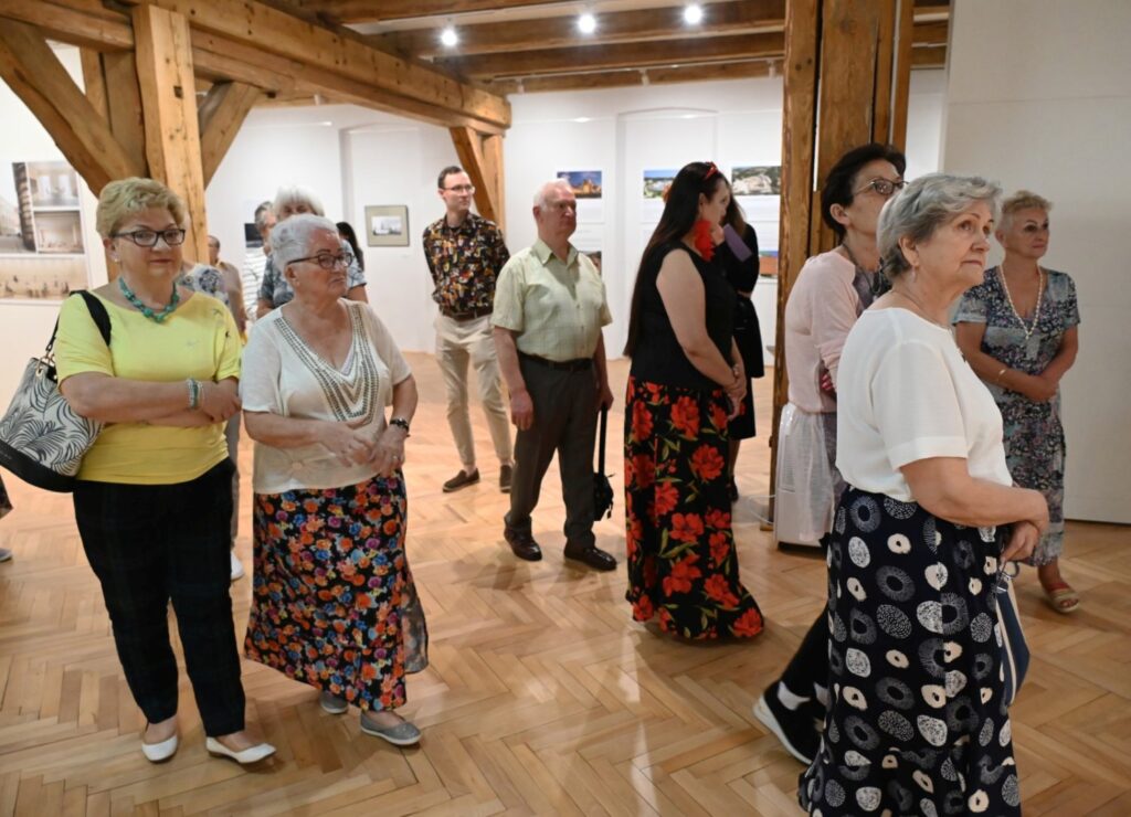 Grupa zwiedzających ogląda wystawę w sali z drewnianymi belkami stropowymi; na ścianach prezentowane są fotografie i materiały dotyczące architektury; w tle widoczne są kolejne części ekspozycji (kliknięcie powiększy zdjęcie).
