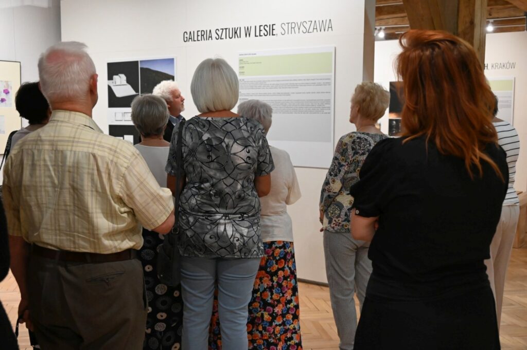Zwiedzający oglądają planszę zatytułowaną „Galeria sztuki w Lesie"; na planszy przedstawiono fotografie i informacje dotyczące ekspozycji; grupa stoi blisko ściany i słucha oprowadzania (kliknięcie powiększy zdjęcie).