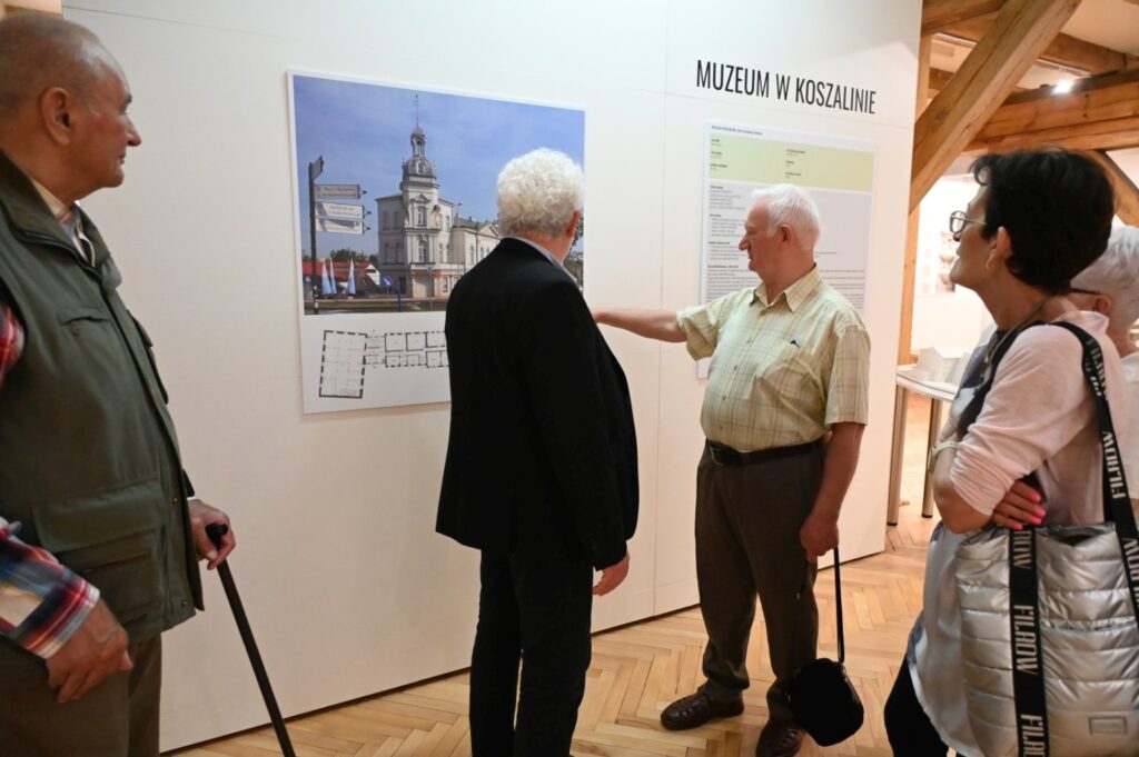 Zwiedzający oglądają planszę wystawy zatytułowaną „Muzeum w Koszalinie”; mężczyzna wskazuje informacje na planszy przedstawiającej fotografię budynku i kalendarium (kliknięcie powiększy zdjęcie).