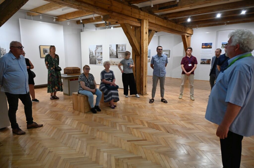 Uczestnicy oprowadzania stoją w przestronnej sali wystawowej; prowadzący rozmawia z grupą, a na ścianach widoczne są fotografie i materiały ekspozycyjne; w głębi znajduje się makieta na postumencie (kliknięcie powiększy zdjęcie).