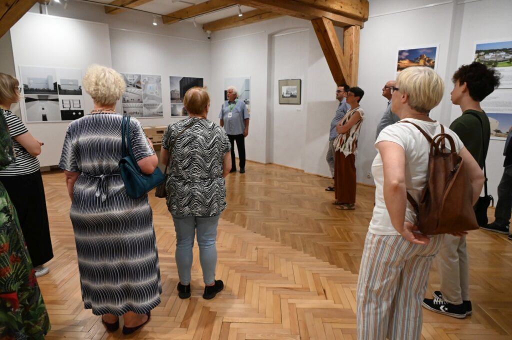 Grupa zwiedzających stoi w sali wystawowej i ogląda fotografie oraz plansze; część osób zwrócona jest w stronę prowadzącego oprowadzanie; w centrum sali ustawiono drewnianą ławkę (kliknięcie powiększy zdjęcie).
