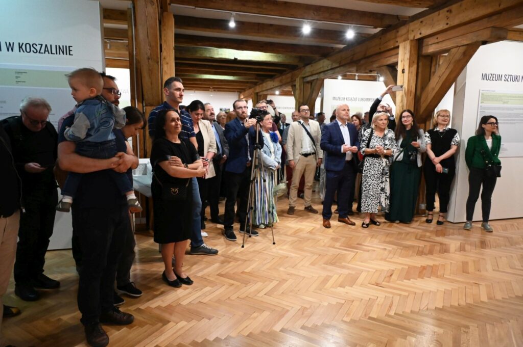 Publiczność zgromadzona w sali muzealnej słucha wystąpienia, stojąc wśród drewnianych elementów konstrukcyjnych wnętrza (kliknięcie powiększy zdjęcie).