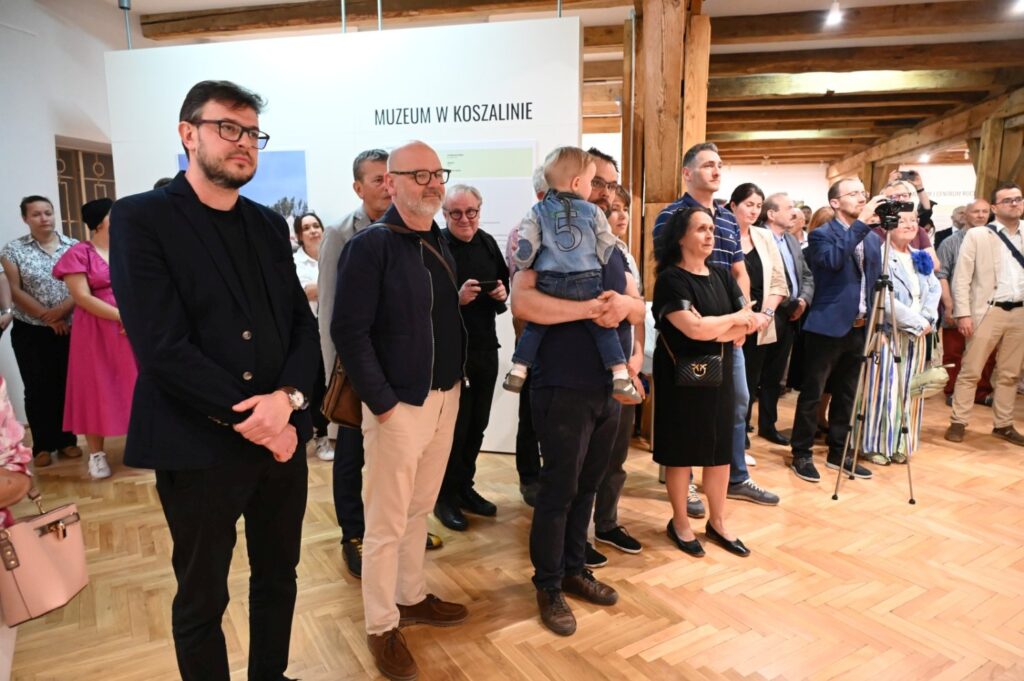 Grupa gości stoi i obserwuje prezentację podczas otwarcia wystawy w muzeum (kliknięcie powiększy zdjęcie).