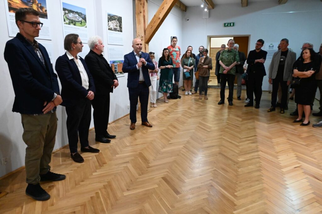 Uczestnicy wernisażu stoją w sali wystawowej, słuchając przemówienia otwierającego wydarzenie (kliknięcie powiększy zdjęcie).