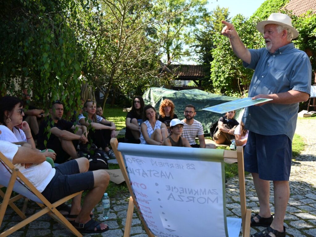 Dyrektor muzeum prowadzi prezentację na świeżym powietrzu, wskazując ręką, przed grupą siedzących uczestników (kliknięcie powiększy zdjęcie).