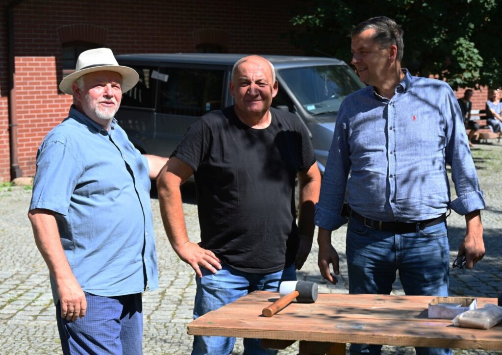 Trzech mężczyzn stoi przy stole z narzędziami na podwórzu, pozując do zdjęcia podczas wydarzenia (kliknięcie powiększy zdjęcie).