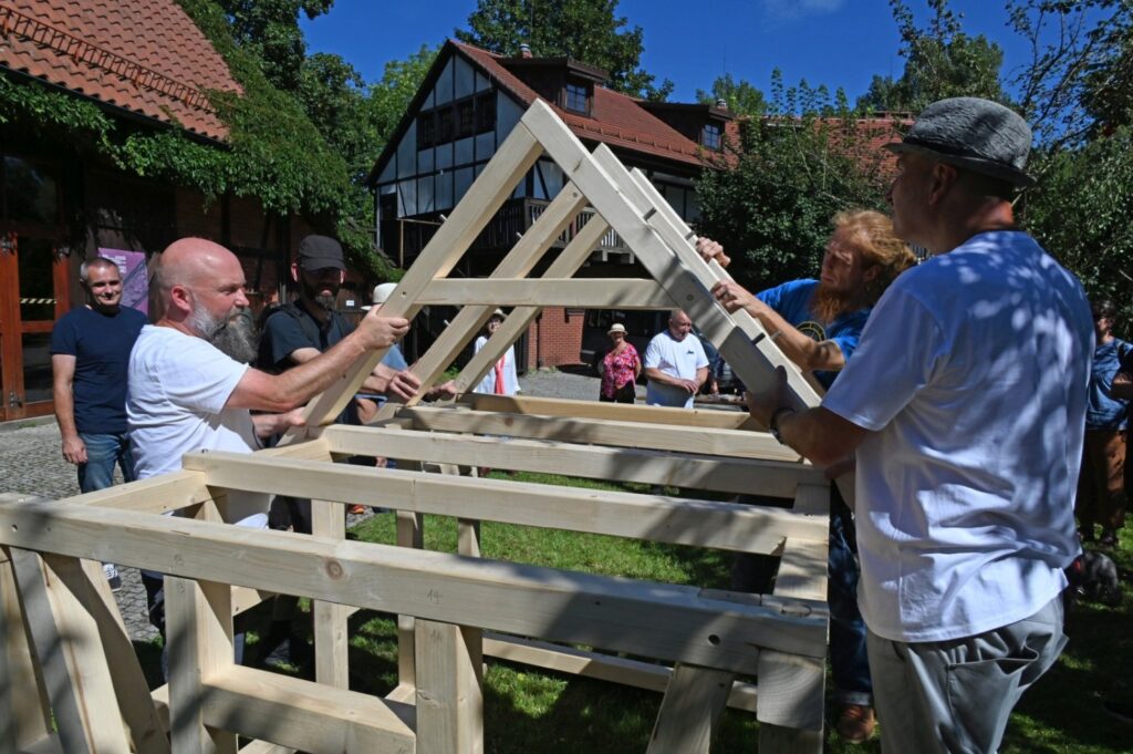 Uczestnicy warsztatów montują drewnianą więźbę dachową na konstrukcji szkieletowej na świeżym powietrzu (kliknięcie powiększy zdjęcie).