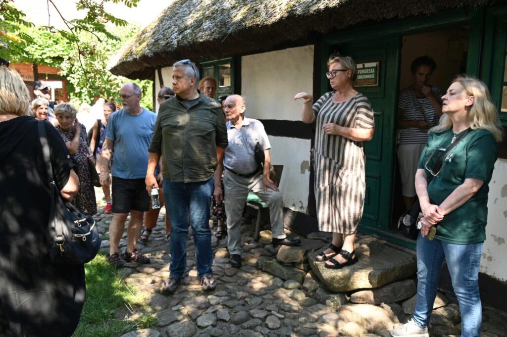Grupa uczestników stoi przed tradycyjnym domem, słuchając przewodnika podczas zwiedzania (kliknięcie powiększy zdjęcie).