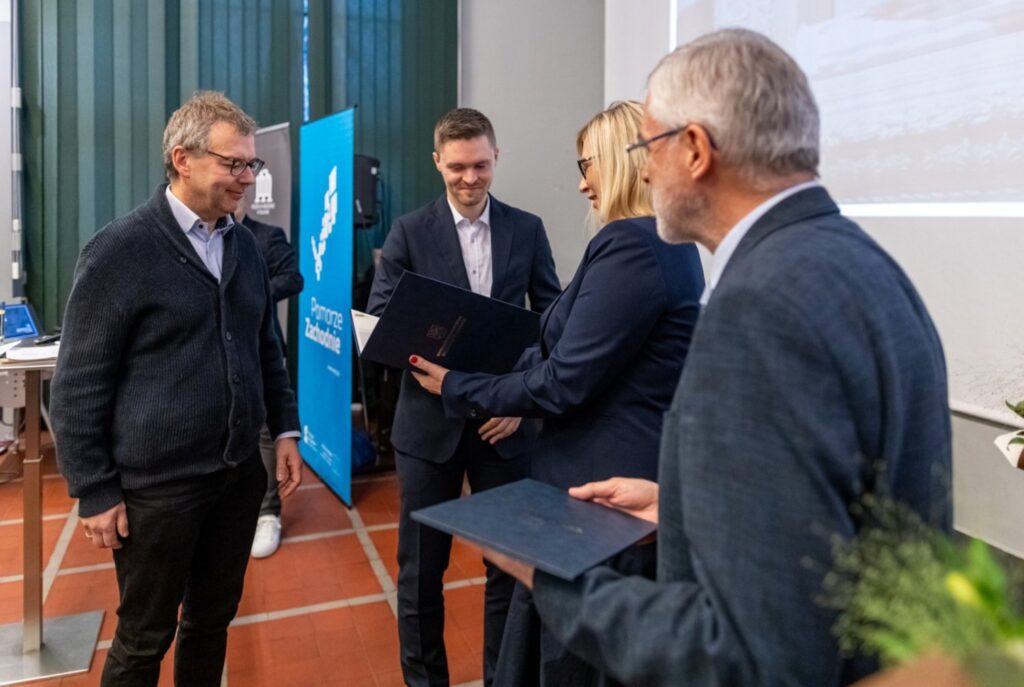 Uczestnicy ceremonii wręczenia nagród przekazują dyplom jednemu z laureatów (kliknięcie powiększy zdjęcie).