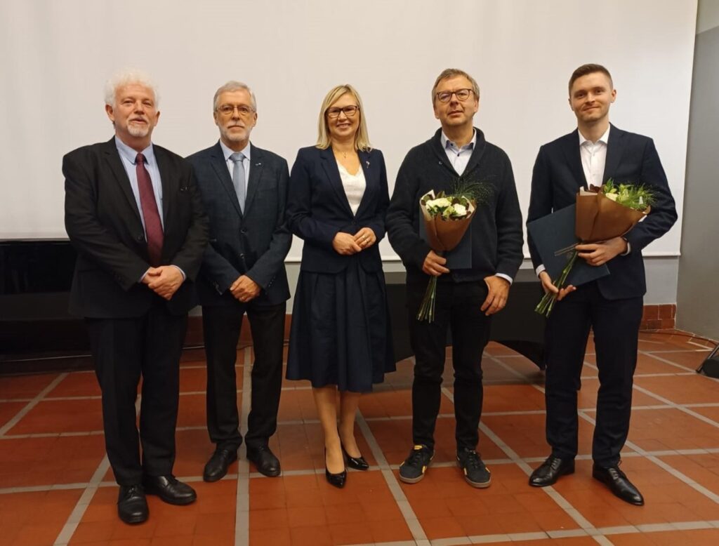 Grupa laureatów stoi razem z organizatorami, trzymając bukiety i dyplomy na scenie (kliknięcie powiększy zdjęcie).