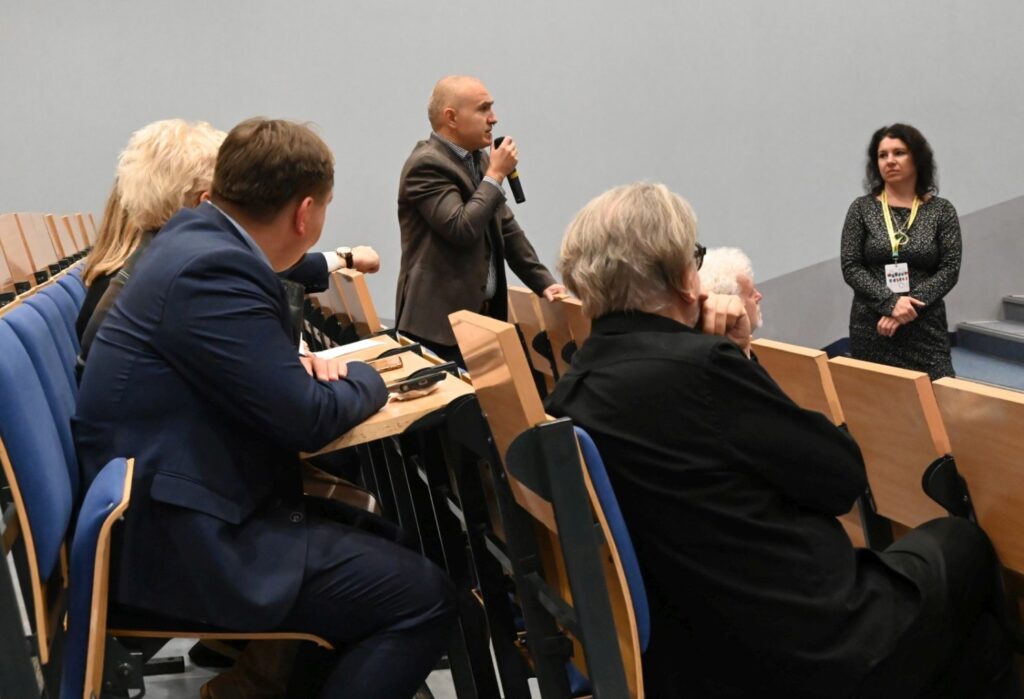 Panel dyskusyjny, uczestnicy siedzą przy stole, a jeden z mówców zabiera głos stojąc (kliknięcie powiększy zdjęcie).