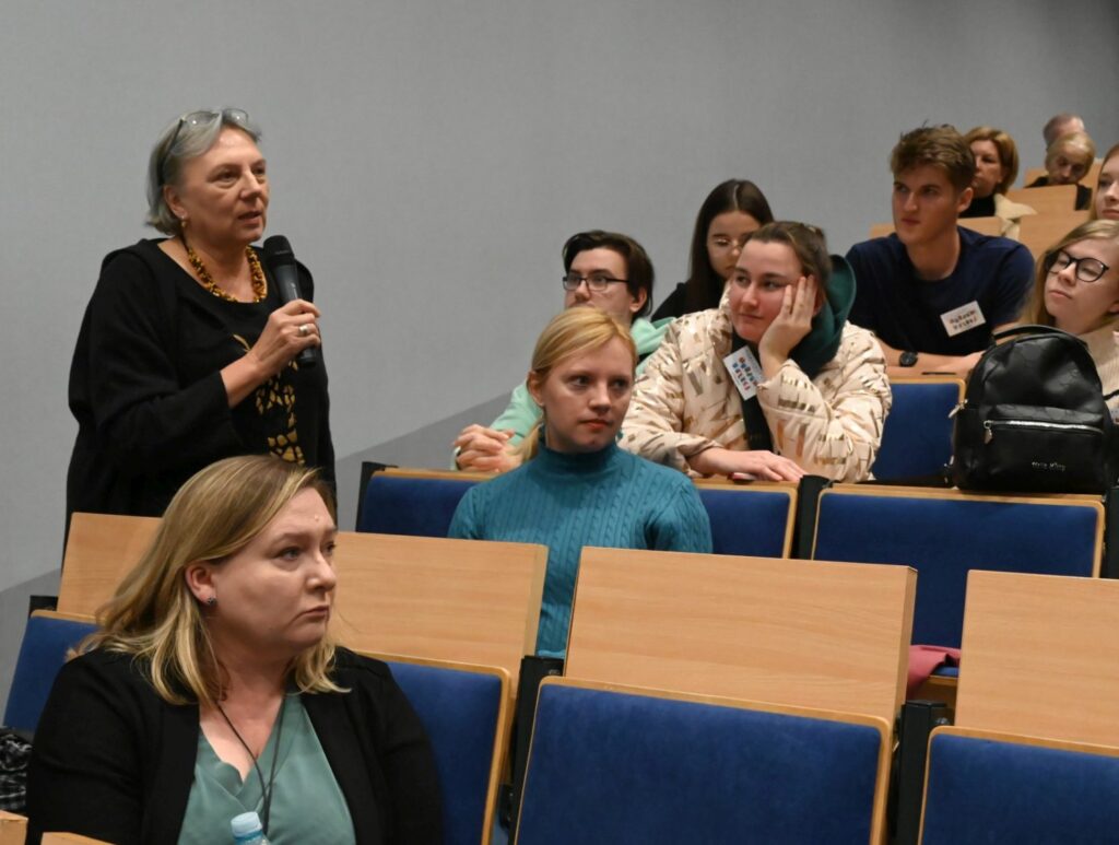 Uczestniczka z widowni zabiera głos, trzymając mikrofon, inni słuchają (kliknięcie powiększy zdjęcie).
