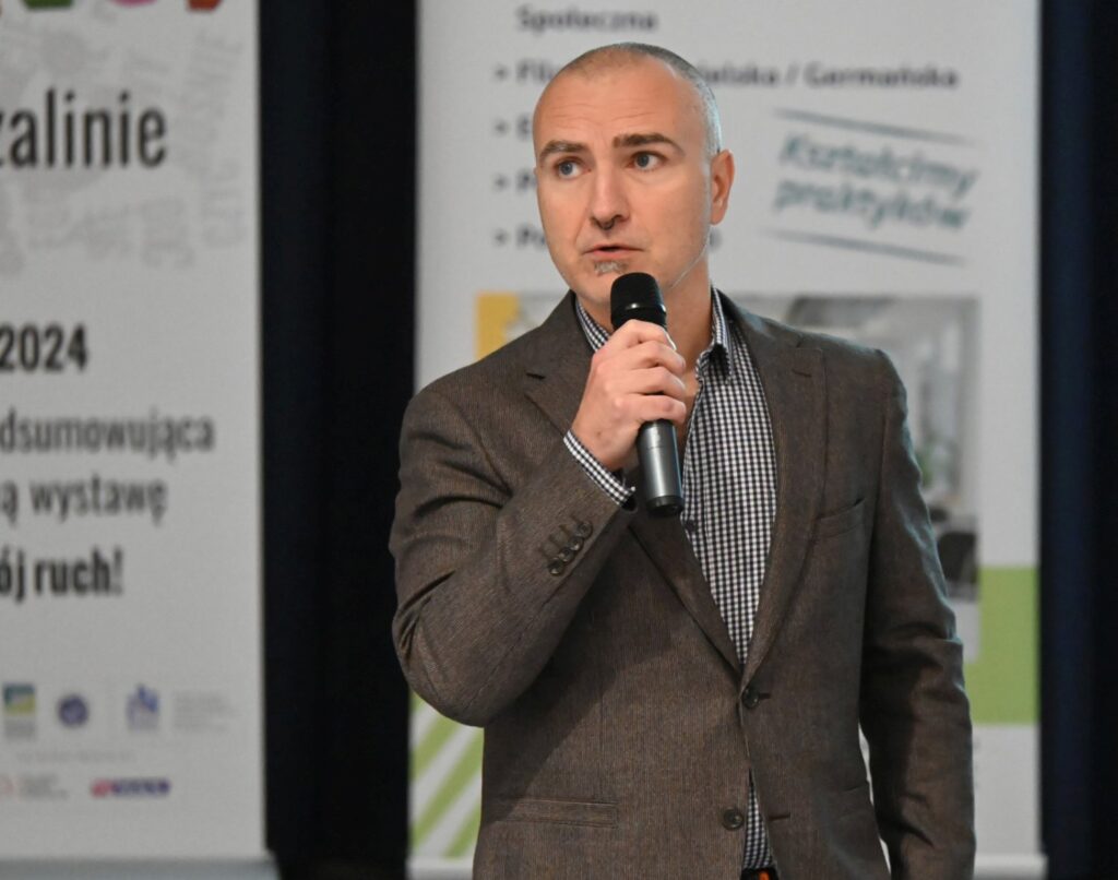 Mężczyzna przemawia do mikrofonu podczas konferencji, stojąc przed banerem wydarzenia (kliknięcie powiększy zdjęcie).