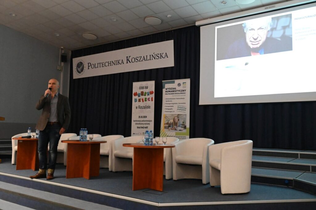 Prelegent stoi na scenie i prowadzi prezentację przed publicznością w sali konferencyjnej (kliknięcie powiększy zdjęcie).