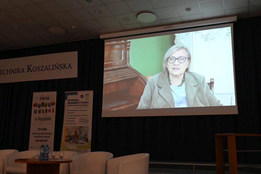 Na ekranie w sali konferencyjnej wyświetlona jest osoba biorąca udział zdalnie w wydarzeniu (kliknięcie powiększy zdjęcie).