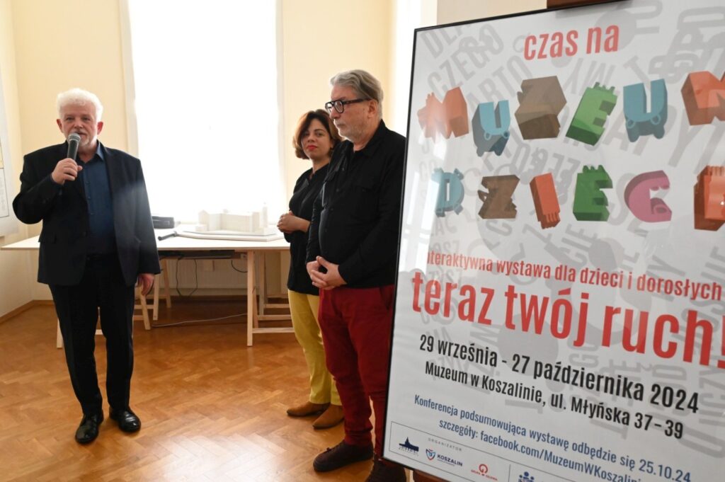 Mężczyzna trzyma mikrofon i przemawia podczas otwarcia wystawy, obok niego stoją dwie osoby; w tle widoczny jest plakat zapowiadający interaktywną wystawę dla dzieci i dorosłych „Czas na Twój Ruch”, prezentowaną w Muzeum w Koszalinie (kliknięcie powiększy zdjęcie).