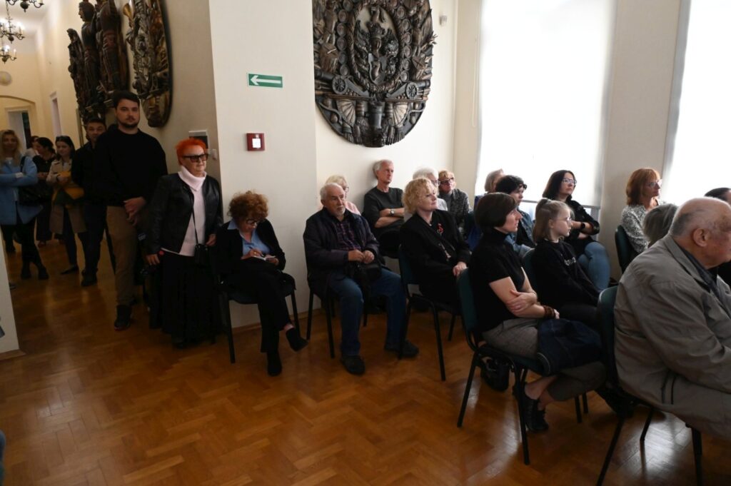 W sali muzealnej siedzi liczna grupa dorosłych i dzieci uczestniczących w otwarciu wystawy; część osób rozmawia i obserwuje wydarzenie, a w tle widoczne są drewniane elementy wnętrza oraz dekoracyjne eksponaty na ścianach (kliknięcie powiększy zdjęcie).