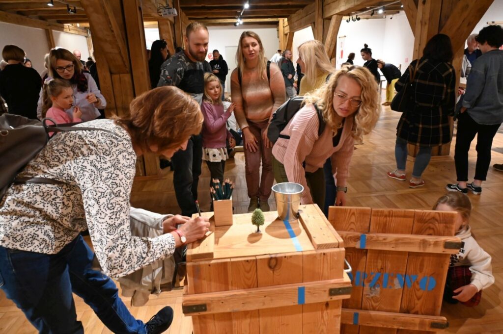 Dorośli i dzieci uczestniczą w interaktywnych zajęciach przy drewnianym stanowisku; kobieta pochyla się nad skrzynią i wykonuje zadanie z przygotowanymi materiałami, podczas gdy pozostali uczestnicy obserwują przebieg zabawy i czekają na swoją kolej (kliknięcie powiększy zdjęcie).