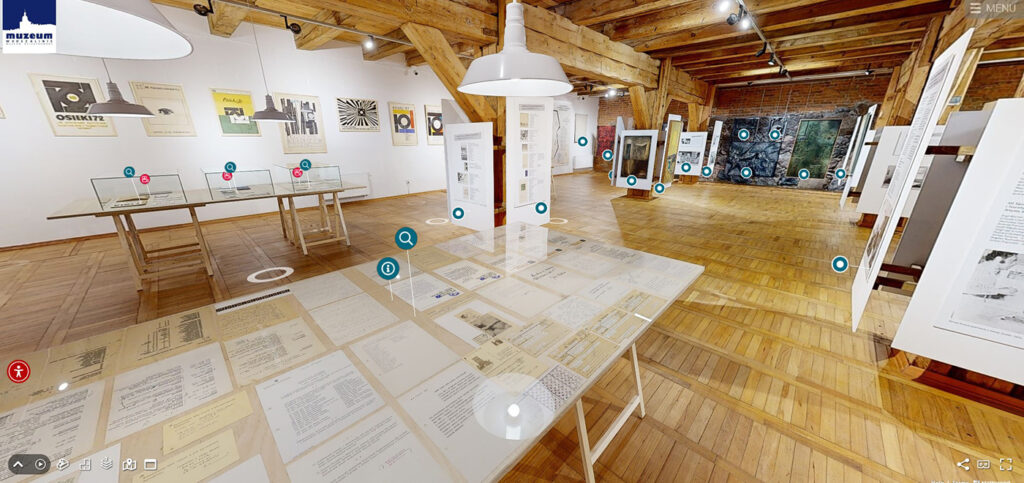 Jasna przestrzeń galerii z drewnianą podłogą i belkowanym sufitem na stołach i ścianach prezentowane grafiki oraz dokumenty a na podłodze plansze ekspozycyjne widoczne znaczniki interaktywne wirtualnego spaceru (kliknięcie powiększy zdjęcie).