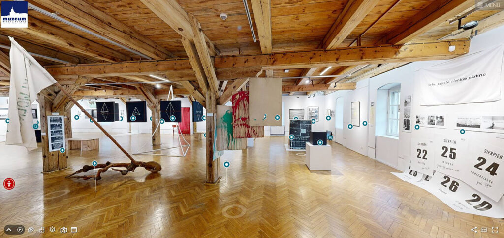 Duża sala wystawowa z drewnianymi belkami i parkietem prezentująca instalacje artystyczne tkaniny oraz plansze informacyjne w tle widoczne stanowiska ekspozycyjne i punkty nawigacyjne spaceru (kliknięcie powiększy zdjęcie).
