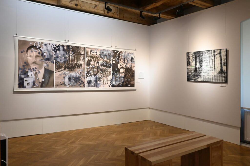 Sala wystawowa z wielkoformatową czarno-białą pracą złożoną z kilku części oraz fotografią przedstawiającą leśny krajobraz; przed ścianą ustawiono drewnianą ławkę (kliknięcie powiększy zdjęcie).