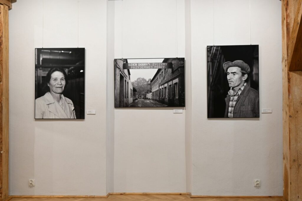 Trzy czarno-białe fotografie przedstawiające portret kobiety, fragment ulicy oraz portret mężczyzny w czapce; prace zawieszono na białej ścianie pomiędzy drewnianymi elementami konstrukcji budynku (kliknięcie powiększy zdjęcie).
