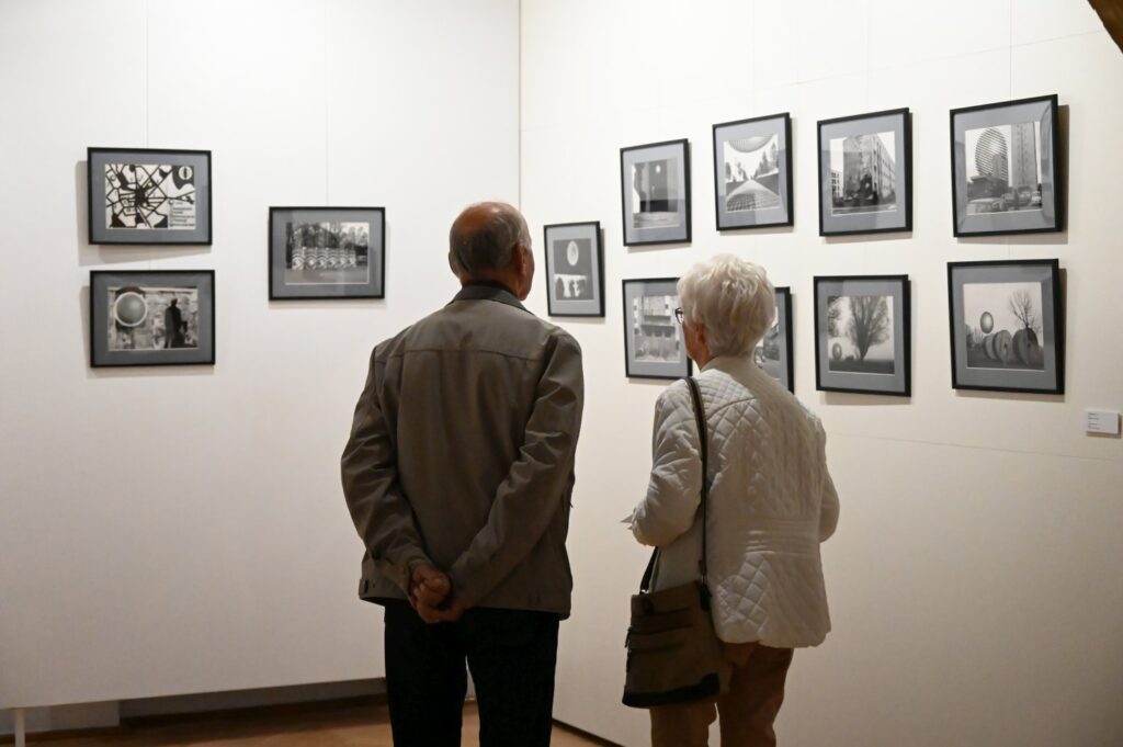 Dwoje zwiedzających ogląda serię czarno-białych fotografii prezentowanych na ścianie galerii; prace są rozmieszczone w kilku rzędach (kliknięcie powiększy zdjęcie).