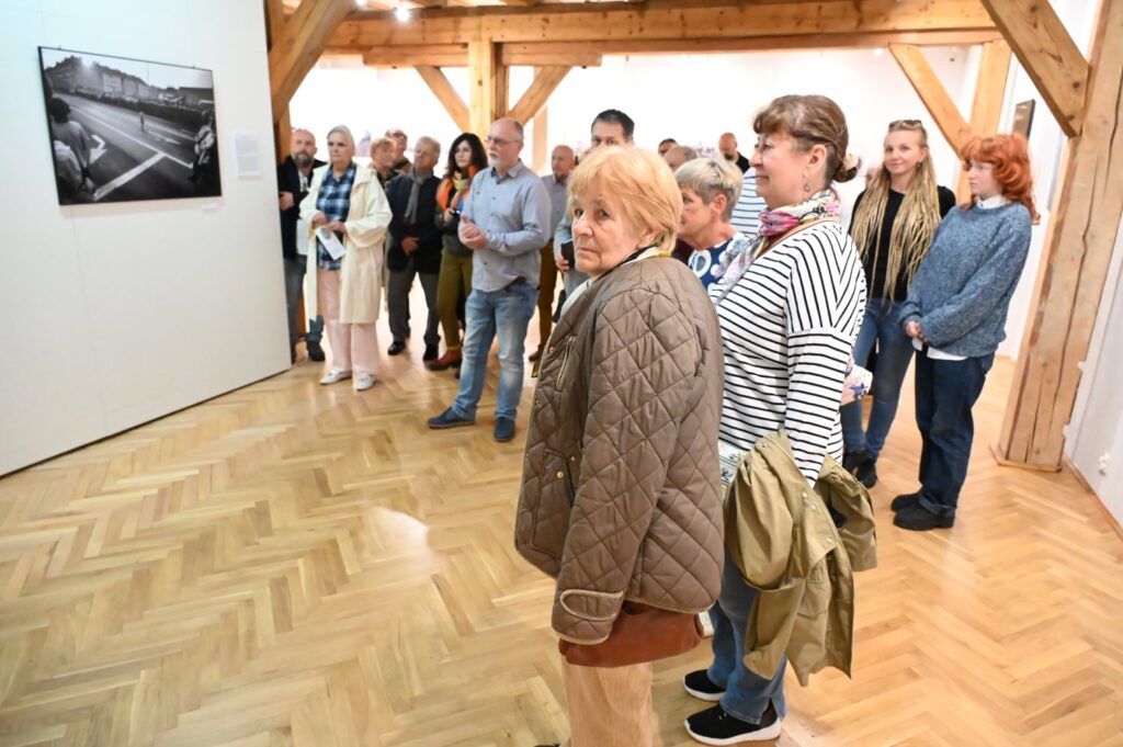 Grupa zwiedzających ogląda fotografie i prace artystyczne prezentowane w sali wystawowej; osoby stoją wzdłuż ściany i słuchają oprowadzania (kliknięcie powiększy zdjęcie).