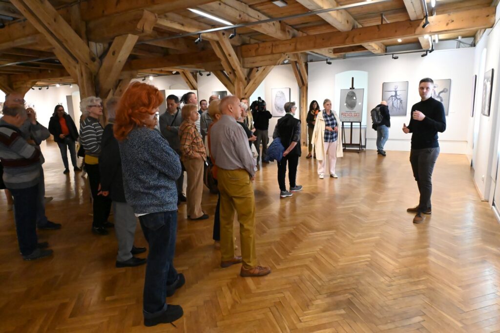 Grupa zwiedzających uczestniczy w oprowadzaniu po wystawie; prowadzący stoi w głębi sali i opowiada o prezentowanych pracach, a wokół znajdują się fotografie i dzieła sztuki (kliknięcie powiększy zdjęcie).