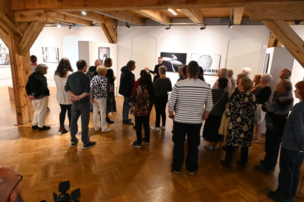 Grupa zwiedzających uczestniczy w oprowadzaniu po wystawie; osoby stoją w sali i oglądają prezentowane na ścianach fotografie oraz dzieła sztuki (kliknięcie powiększy zdjęcie).