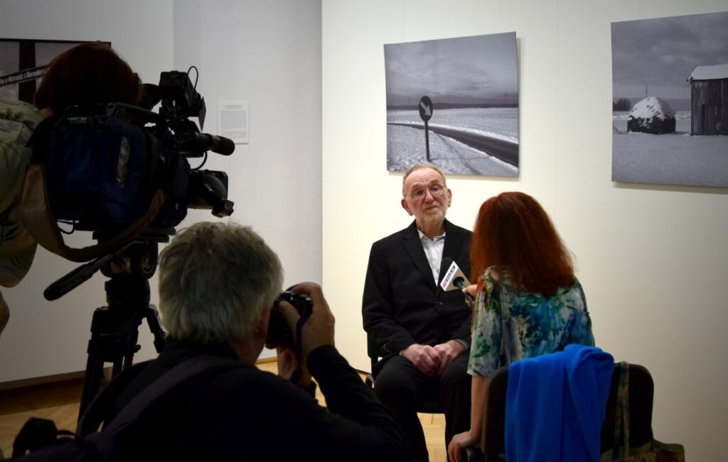 Wywiad podczas wydarzenia; mężczyzna siedzi na krześle przed mikrofonem, a obok niego znajduje się dziennikarka, w tle widoczne są czarno-białe fotografie (kliknięcie powiększy zdjęcie).