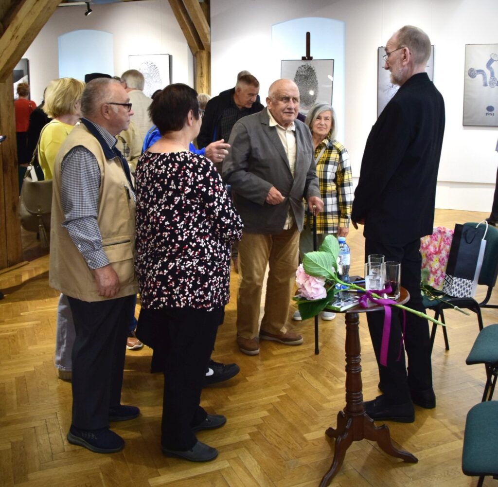 Rozmowa uczestników podczas wydarzenia; kilka osób stoi w galerii przy stoliku z kwiatami i napojami, w tle widoczna jest ekspozycja prac artystycznych oraz zgromadzona publiczność (kliknięcie powiększy zdjęcie).