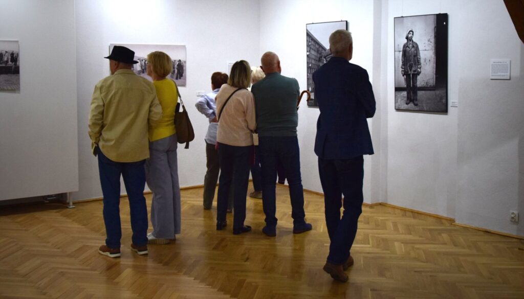 Zwiedzający oglądają wystawę podczas wydarzenia; grupa osób stoi przed pracami artystycznymi rozmieszczonymi na białych ścianach galerii (kliknięcie powiększy zdjęcie).