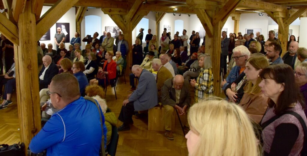 Publiczność podczas wydarzenia w sali wystawowej; liczna grupa osób siedzi i stoi pod drewnianą konstrukcją galerii, część uczestników zwrócona jest w stronę miejsca spotkania (kliknięcie powiększy zdjęcie).