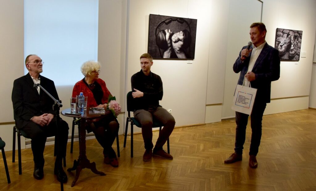 Spotkanie z artystami podczas wystawy; trzy osoby siedzą przy stoliku, a mężczyzna stojący obok trzyma mikrofon i prowadzi rozmowę, w tle widoczne są czarno-białe prace artystyczne (kliknięcie powiększy zdjęcie).