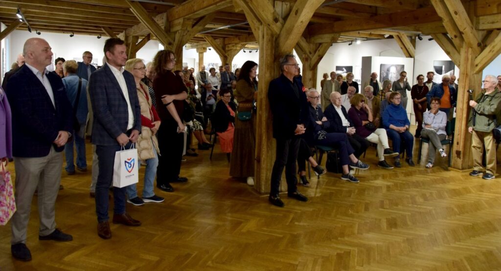 Publiczność podczas spotkania w galerii; część osób stoi, a część siedzi pod drewnianymi belkami sali, wśród uczestników widoczne są osoby prowadzące wydarzenie (kliknięcie powiększy zdjęcie).