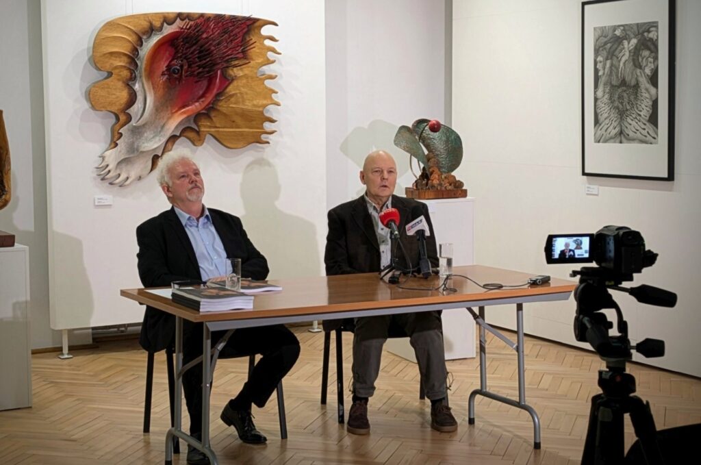 Dwóch mężczyzn siedzi przy stole podczas konferencji prasowej w galerii sztuki, przed nimi mikrofony i kamera nagrywająca (kliknięcie powiększy zdjęcie).
