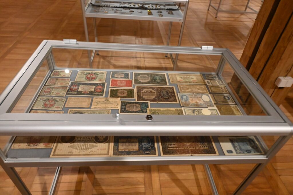 Przeszklona gablota z kolekcją historycznych banknotów i papierowych środków płatniczych; eksponaty ułożono w uporządkowanych rzędach i grupach, prezentując różne wzory, nominały i emisje; wśród materiałów widoczne są również kolorowe druki oraz banknoty o zróżnicowanej szacie graficznej (kliknięcie powiększy zdjęcie).